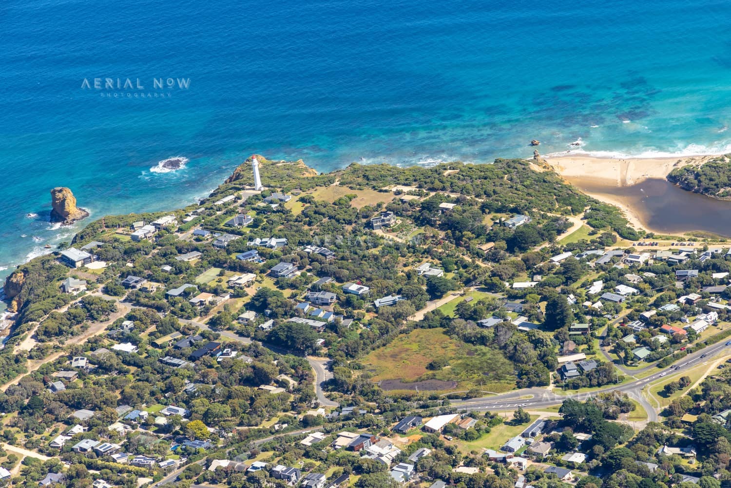 Aerial view of Split Point Lighthouse and Eagle Rock at Aireys Inlet, showing coastal homes, turquoise ocean, beaches and the nearby creek estuary.