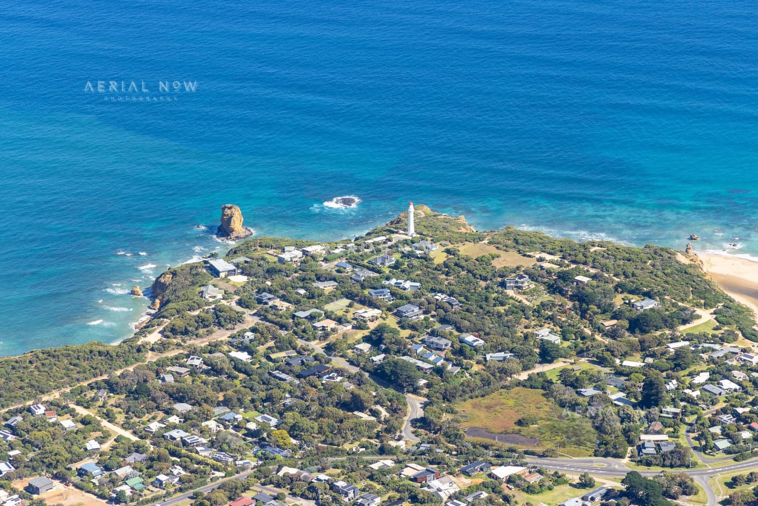 Aerial view of Split Point Lighthouse and Eagle Rock on the Aireys Inlet coastline, with coastal homes, roads, beach and blue ocean surrounding the headland.