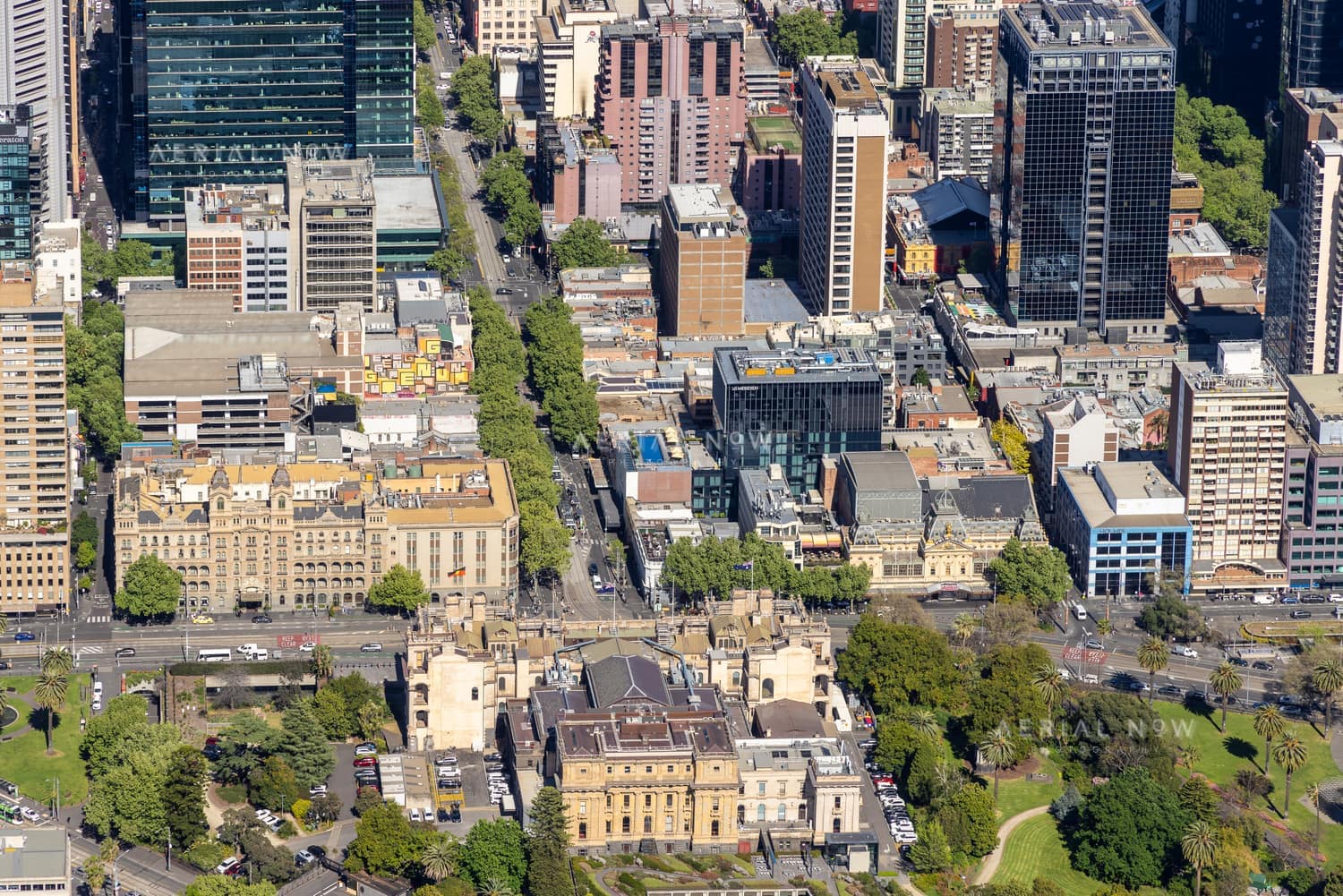 Aerial view of Parliament House beside Spring Street in Melbourne, with surrounding CBD office towers, traffic, city streets and parkland.