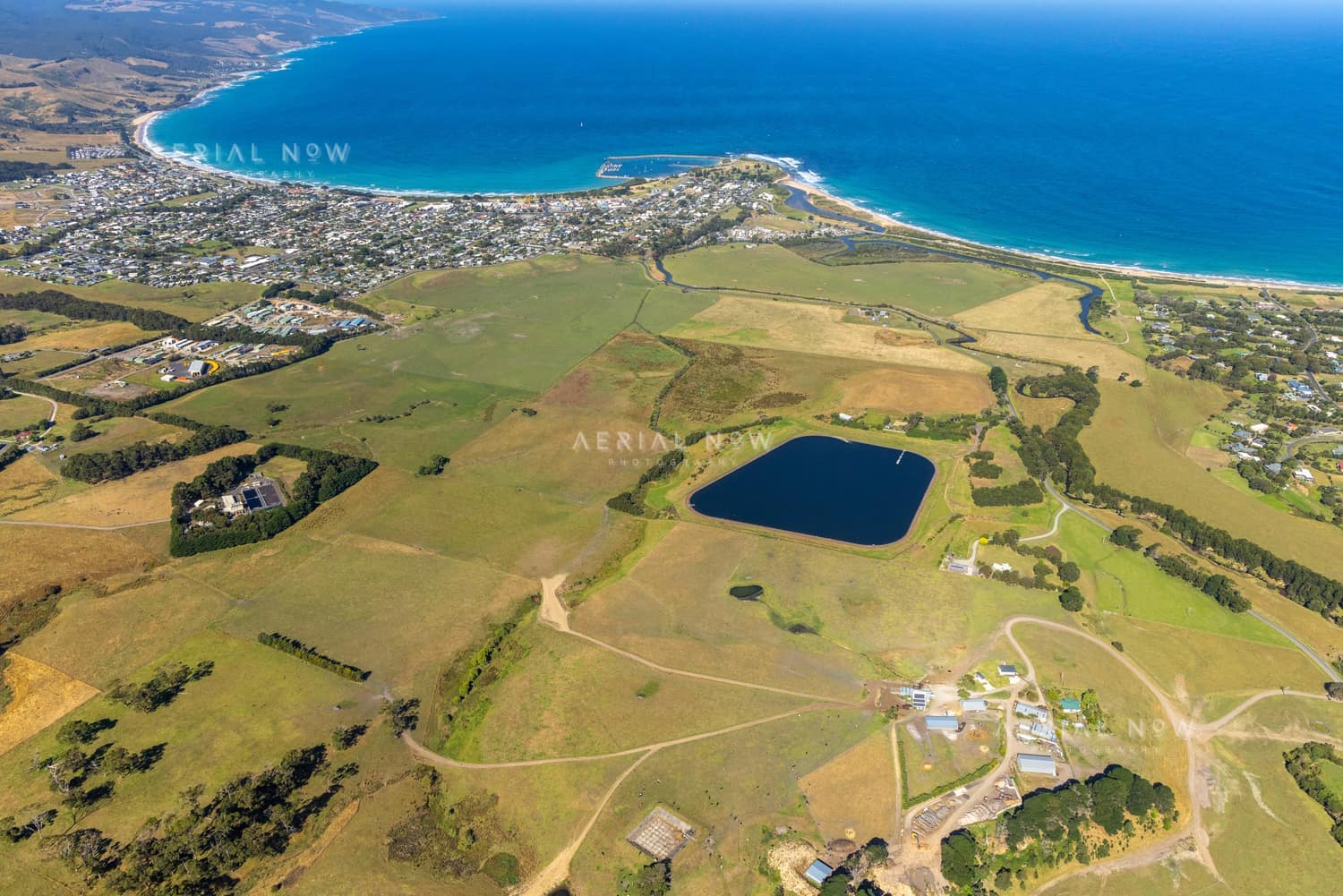 Aerial view of Apollo Bay township and harbour beside the curved coastline, with farmland, a water storage pond and residential areas in Colac Otway, Victoria.