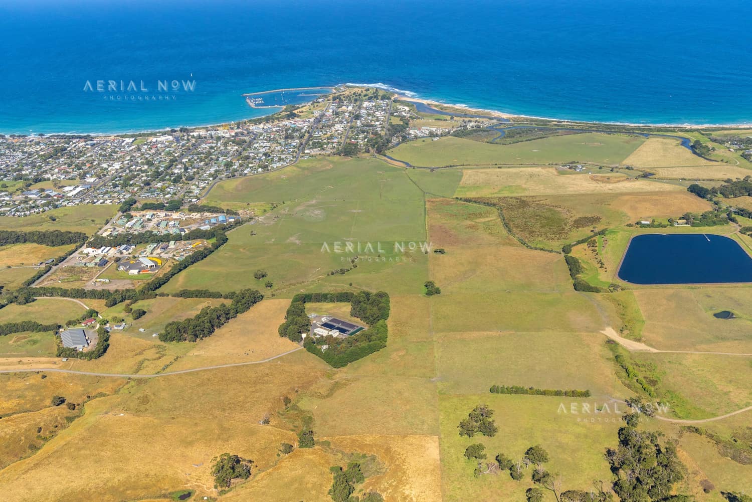 Aerial view of Apollo Bay township and coastline in Victoria, with farmland, rural properties, a farm dam, beaches and blue ocean around the harbour.