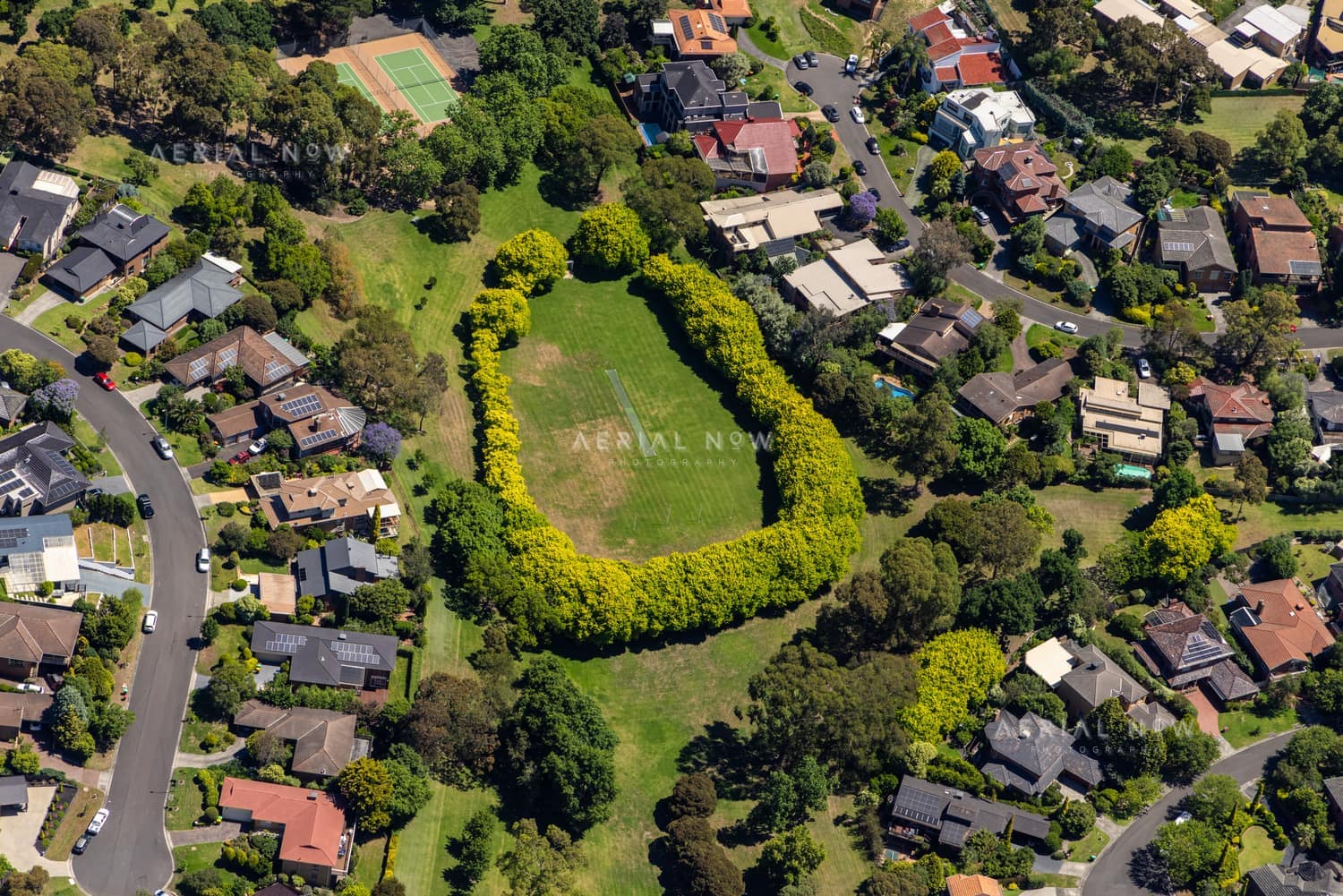 Aerial view of green suburban parkland with a tennis court, surrounding homes and tree-lined streets in Doncaster East, Victoria.