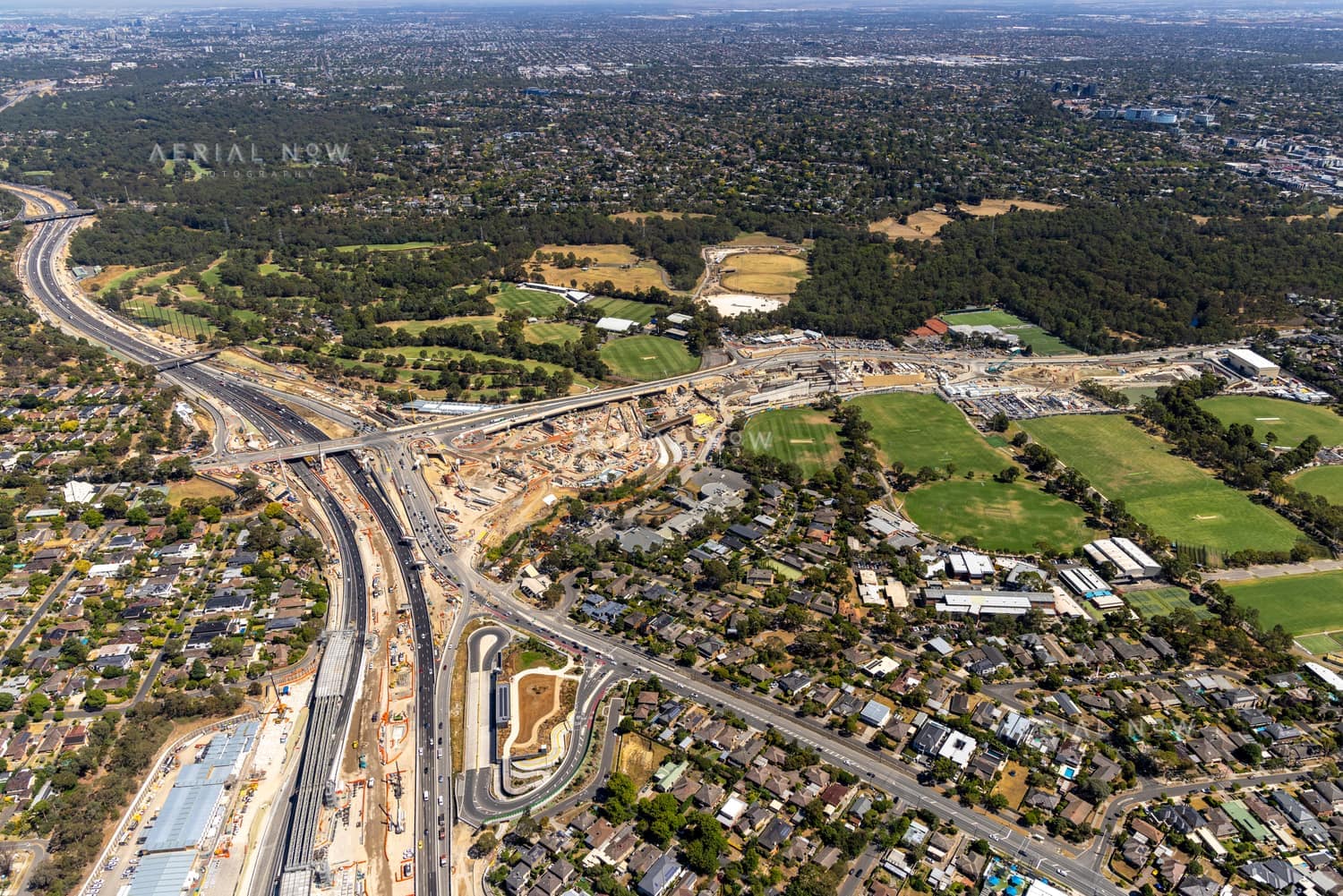 Aerial view of North East Link construction works in Bulleen, showing major road corridors, earthworks, sporting fields, parkland and surrounding suburban streets.