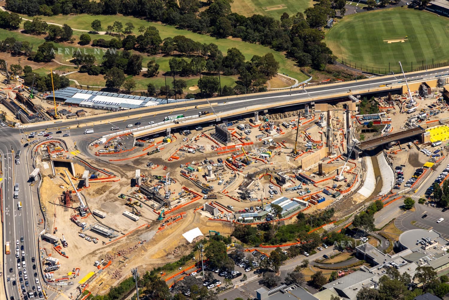 Aerial view of the North East Link construction site beside Manningham Road in Bulleen, with cranes, bridge works, traffic, parkland and a nearby sports oval visible.