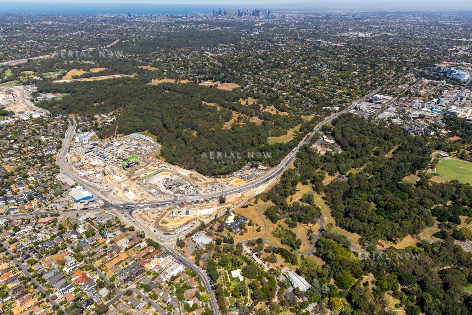 Aerial view of North East Link construction works in Bulleen, with roadworks, surrounding suburban streets, parkland and the Melbourne skyline in the distance.