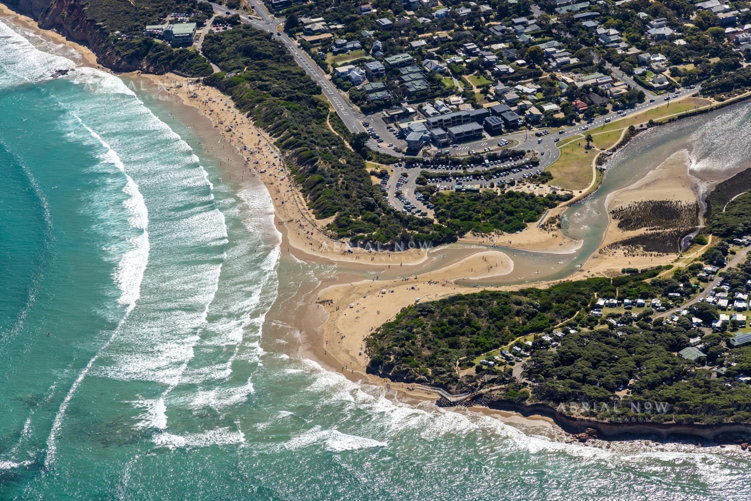 Aerial view of the Anglesea River mouth meeting the surf at Anglesea Beach, with Great Ocean Road, township streets, car parks and coastal vegetation in Anglesea, Victoria.