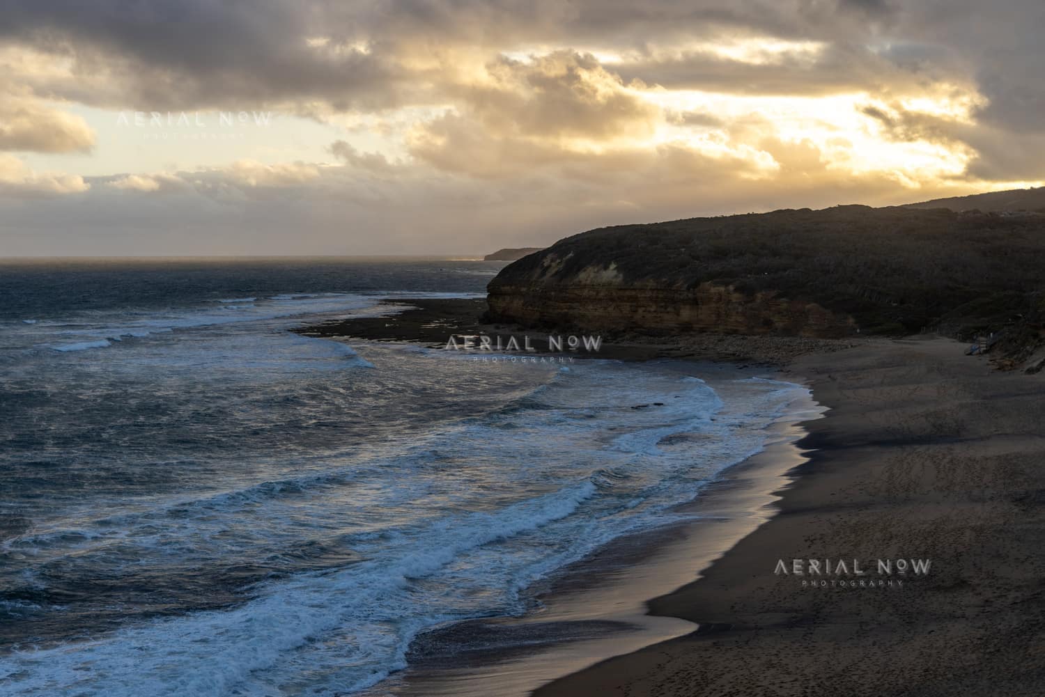 Aerial view of Bells Beach coastline in Bells Beach VIC, showing surf rolling onto the beach below coastal cliffs under a cloudy golden hour sky.