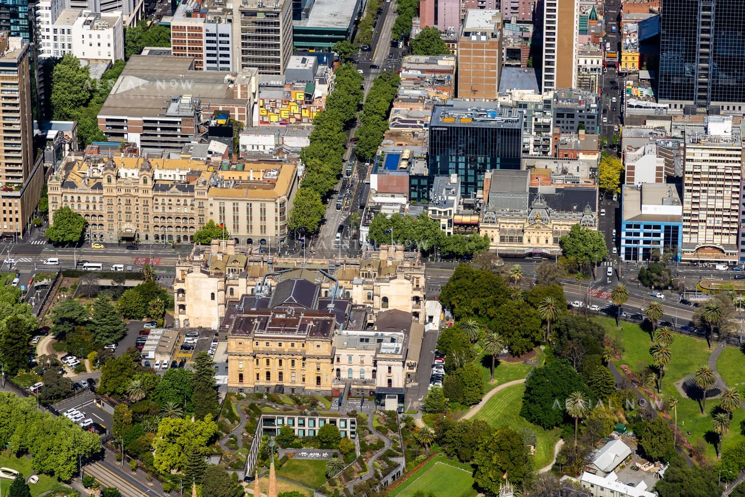 Aerial view of Parliament House beside Treasury Gardens in Melbourne, with The Hotel Windsor, Spring Street traffic, parkland and surrounding CBD buildings visible.