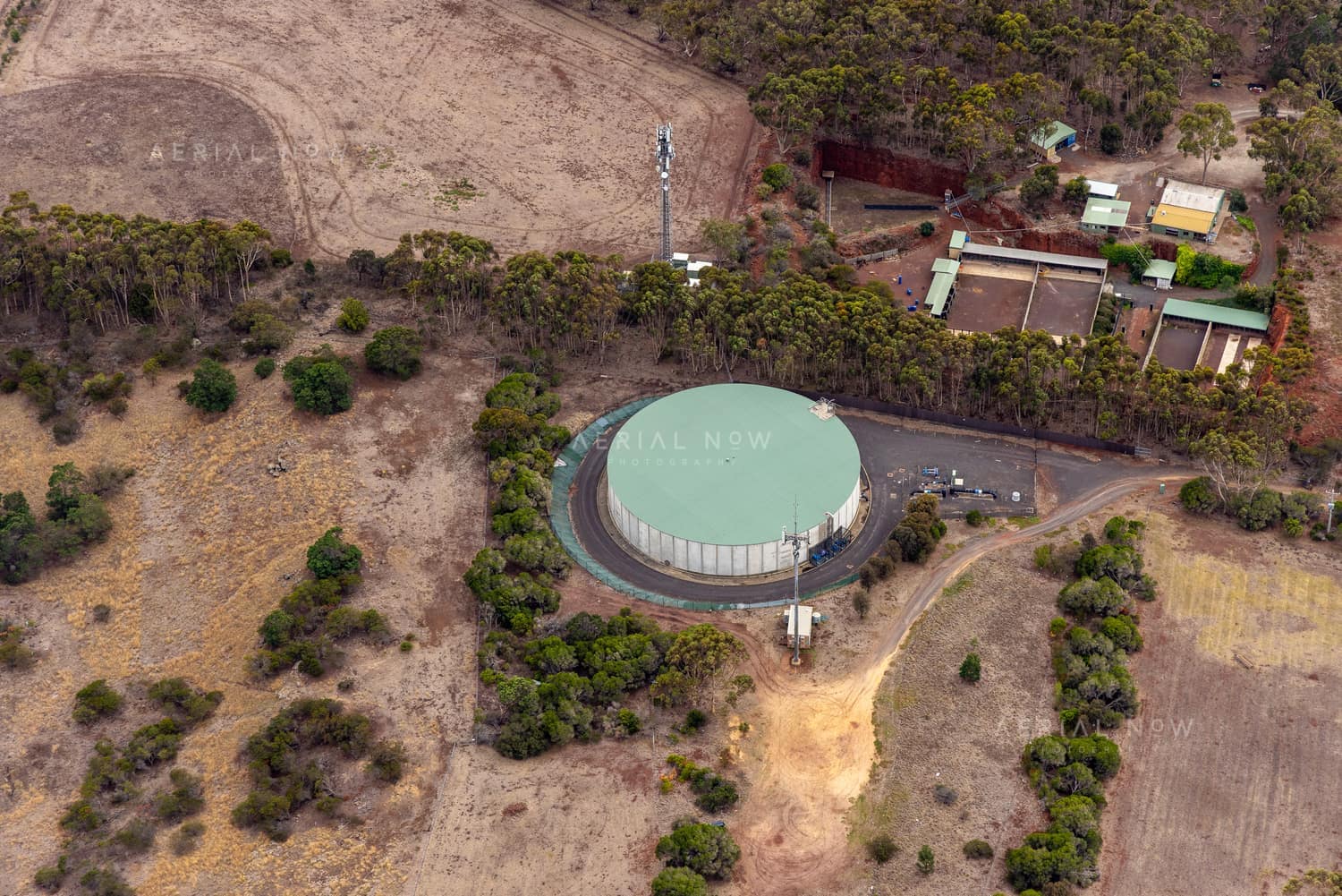 Aerial view of a large circular water storage tank surrounded by dry paddocks, bushland and utility buildings in Mount Duneed, Victoria.