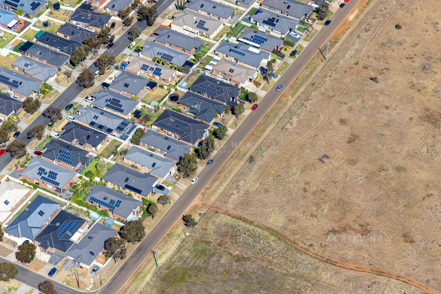 Aerial view of a Brookfield residential estate with solar-panelled houses beside a road and adjoining vacant dry grassland in Victoria.