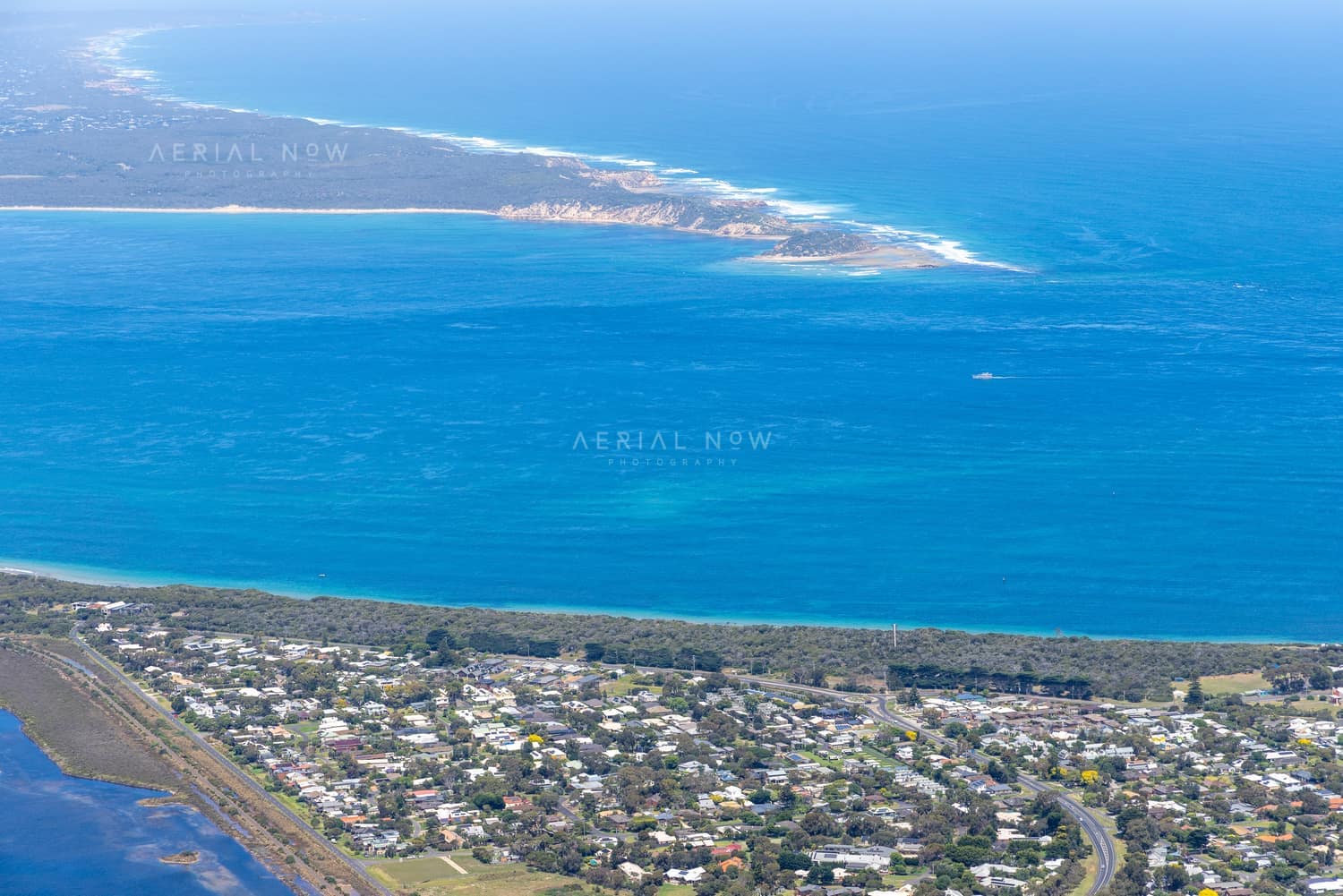 Aerial view of Point Lonsdale residential streets and coastal vegetation beside blue water, with Point Nepean visible across Port Phillip Bay.
