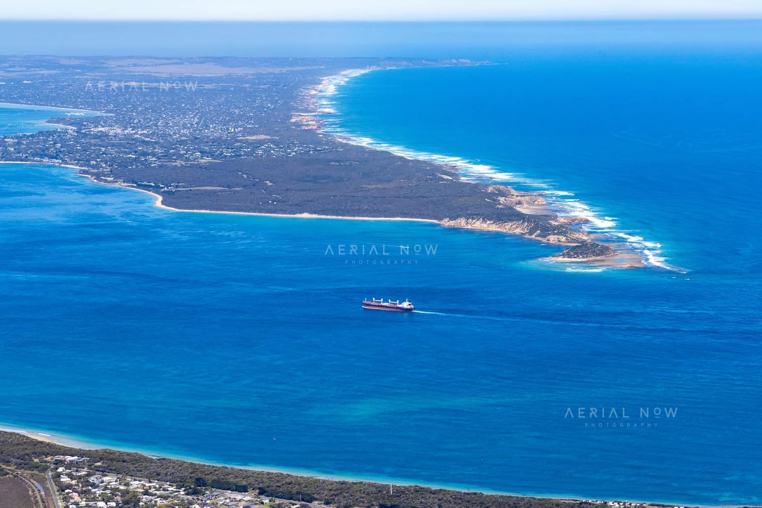 Aerial view of Port Phillip Heads and Point Nepean from Point Lonsdale, with a cargo ship crossing blue water between the coastline and surf beaches.