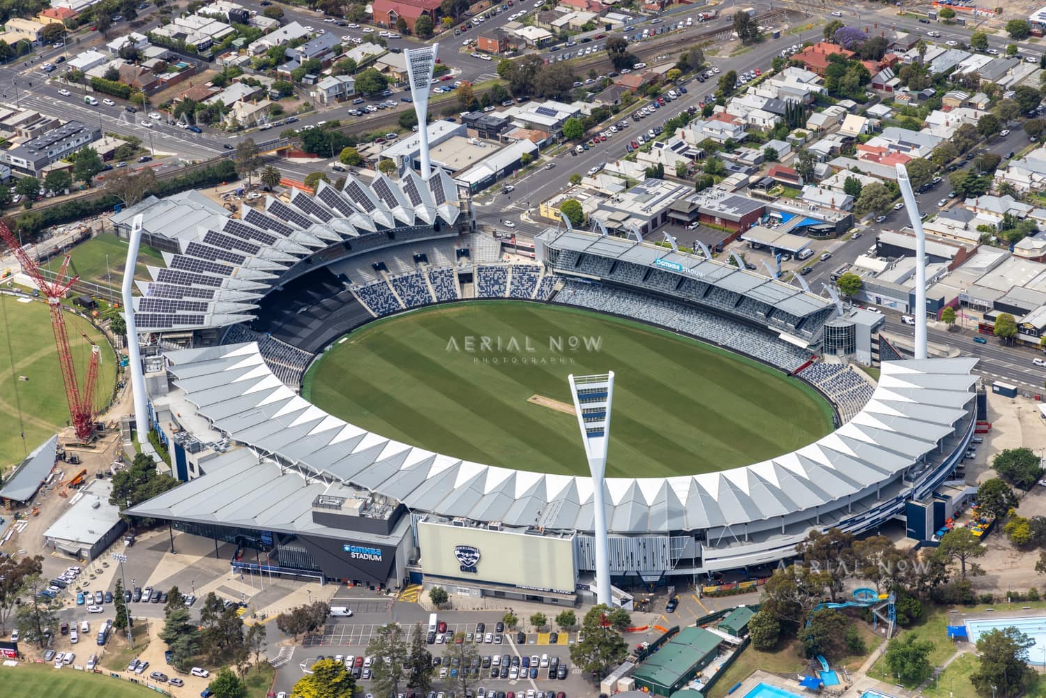 Aerial view of GMHBA Stadium in Kardinia Park, South Geelong, showing the oval, grandstands, roof structures, light towers, nearby streets, car parks and a construction crane.