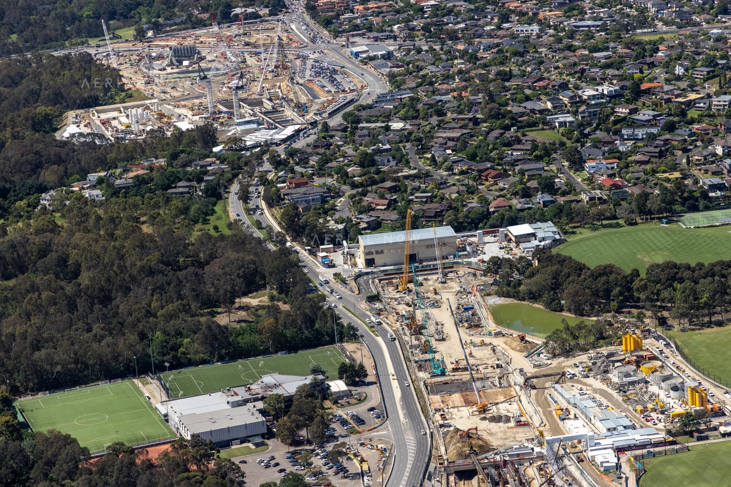 Aerial view of North East Link construction works along Bulleen Road in Bulleen, with cranes, work compounds, sports fields, bushland and surrounding residential streets visible.
