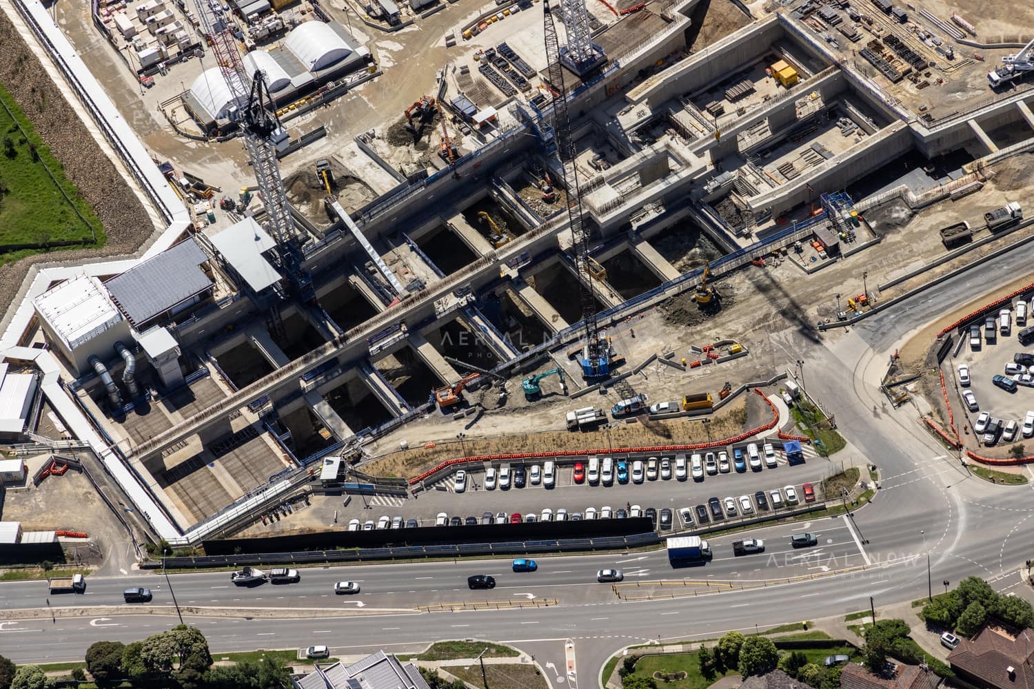 Aerial view of North East Link construction works in Bulleen, showing concrete tunnel and road structures, cranes, work zones, parked vehicles and traffic on the adjoining arterial road.