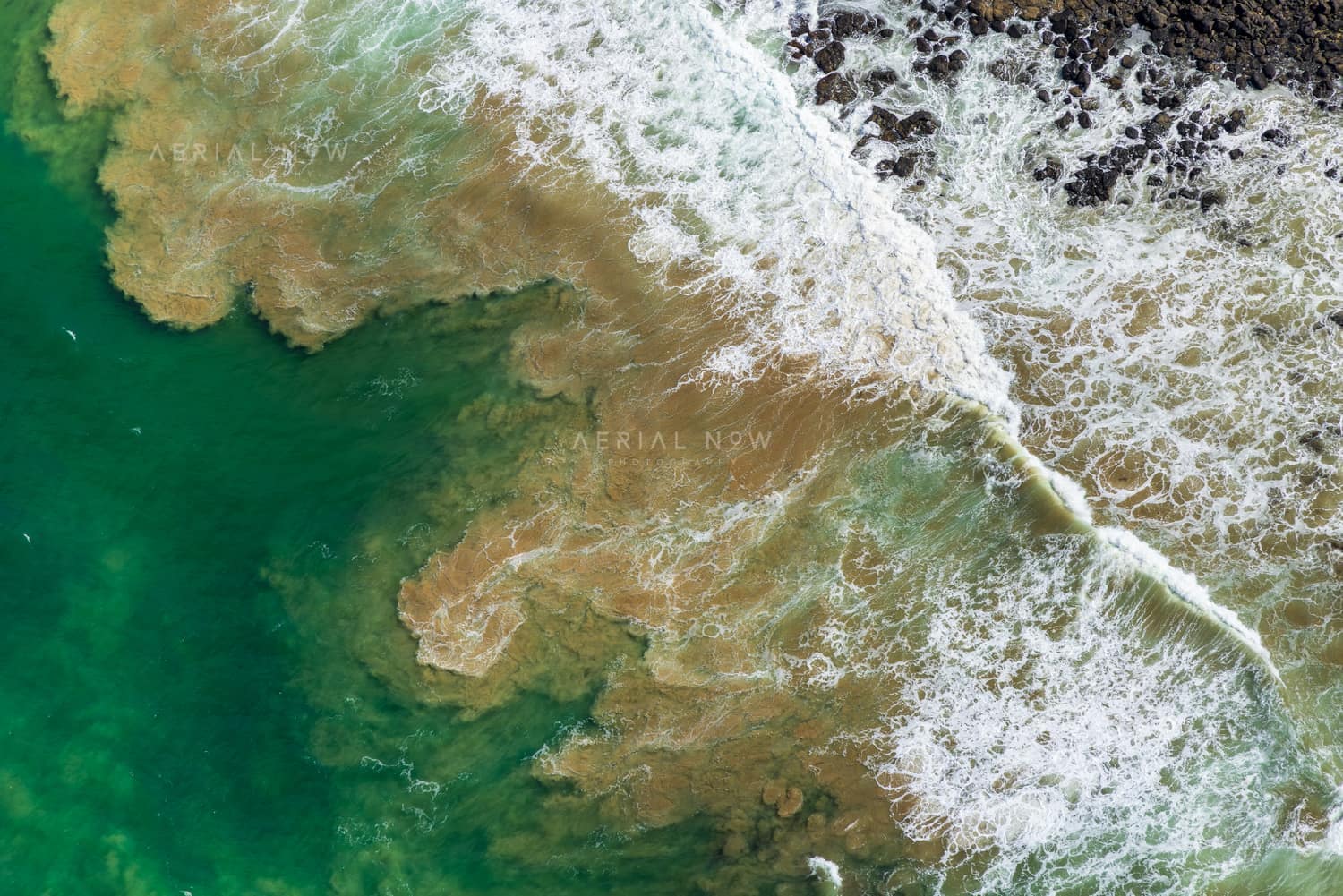 Aerial view of waves breaking over a shallow rocky reef and green coastal water at Breamlea, Victoria.