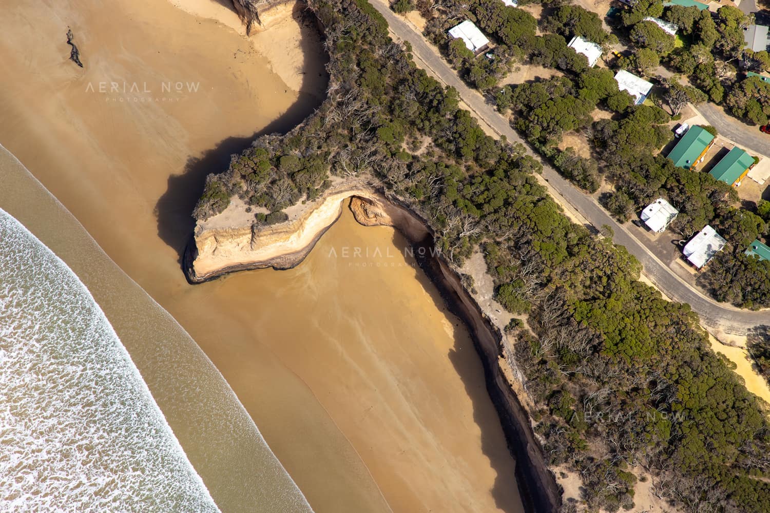 Aerial view of a sandy beach, eroded coastal cliffs, bushland and nearby houses along the Anglesea coastline in Victoria.