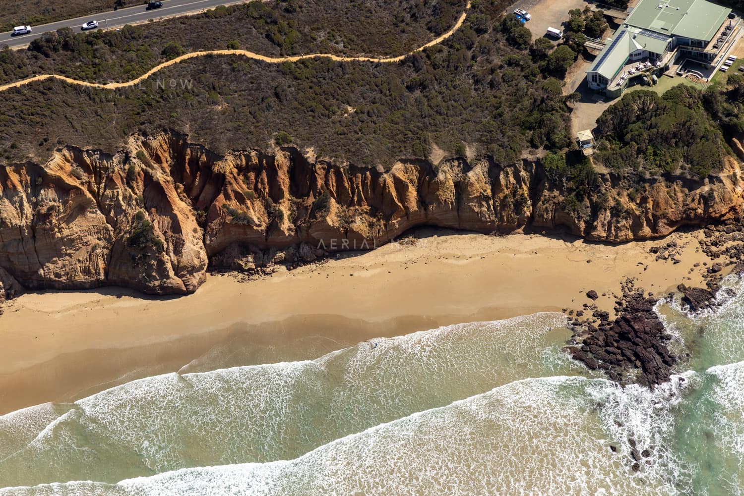 Aerial view of Demons Bluff Beach at Anglesea VIC, showing waves washing onto a narrow sandy beach below eroded sandstone cliffs, with clifftop homes and a coastal road above.