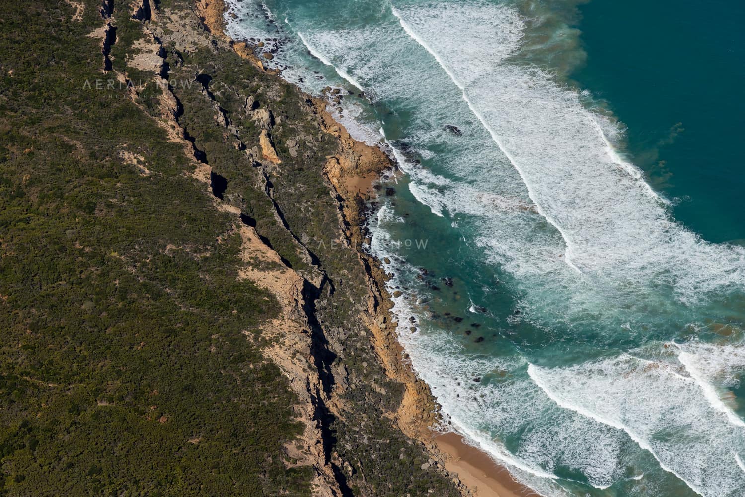 Aerial view of rugged cliffs, coastal scrub, surf and a narrow sandy beach in Port Campbell National Park near Princetown, Victoria.
