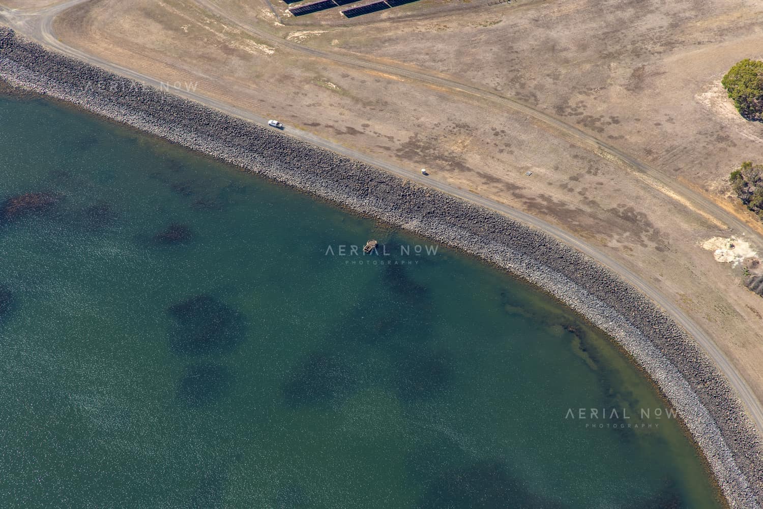 Aerial view of Wurdee Boluc Reservoir shoreline showing green water, a rock-armoured embankment, dry access tracks and vehicles near Wurdiboluc, Victoria.