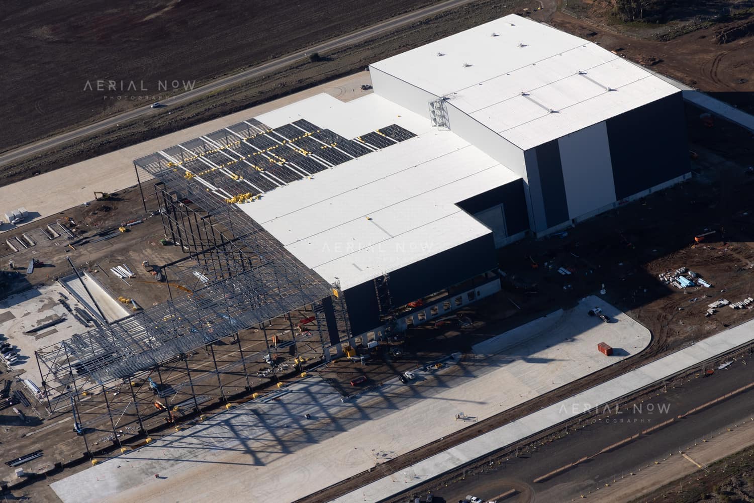 Aerial view of a large industrial warehouse construction site on Brinbrook Street in Tarneit VIC, with white roof panels, steel framing, concrete hardstand and surrounding earthworks.