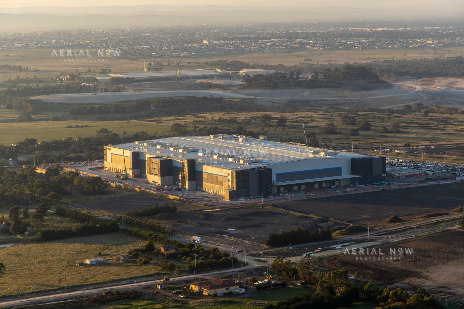 Aerial view of a large industrial warehouse complex surrounded by roads, car parks, open land and developing estates in Mickleham, Victoria.