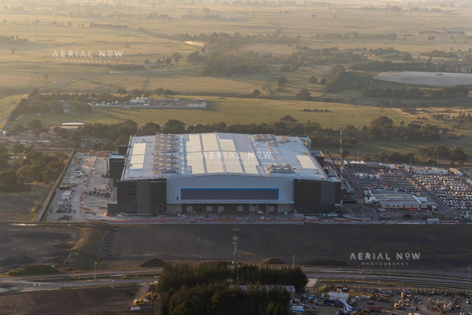 Aerial view of the Amazon Robotics Fulfilment Centre under construction in Craigieburn, with a large warehouse building, loading areas, staff parking and surrounding rural land.