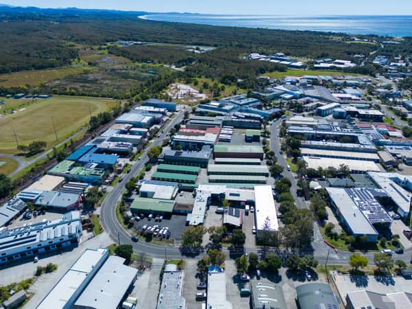 Aerial view of the Centennial Circuit industrial estate in Byron Bay, with warehouses, commercial buildings, parked vehicles, surrounding bushland and the coastline in the background.
