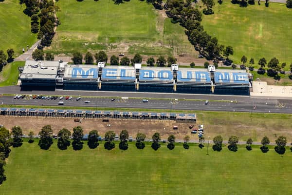 Aerial view of the Albert Park Circuit pit building beside Aughtie Drive in Albert Park, with MELBOURNE roof lettering, parked cars and surrounding sports fields.