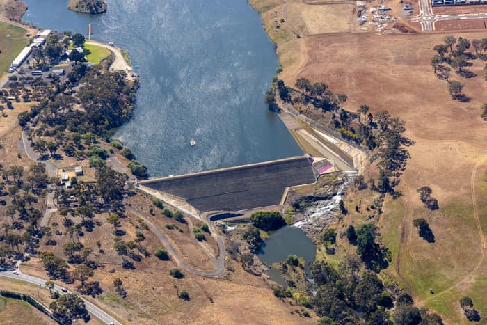 Aerial view of the Melton Reservoir dam wall and spillway near Mount Cottrell Road in Melton, Victoria, with surrounding dry farmland, trees and nearby development.