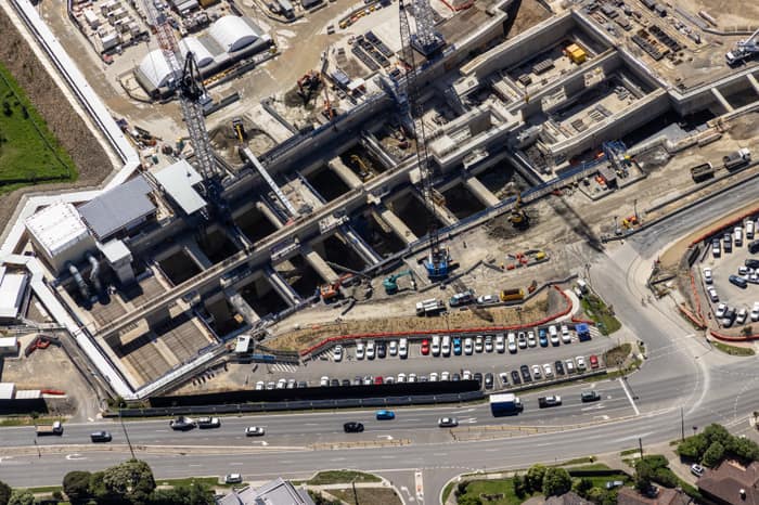 Aerial view of North East Link construction works in Bulleen, showing concrete tunnel and road structures, cranes, work zones, parked vehicles and traffic on the adjoining arterial road.