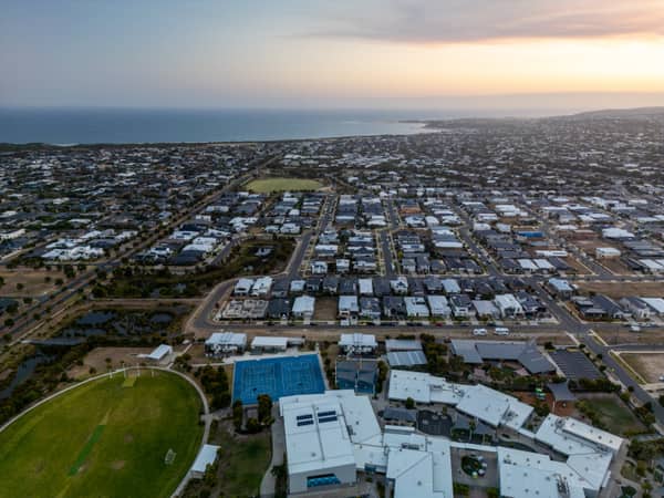 Aerial view over the Quay Boulevard residential area in Torquay VIC, showing suburban streets, sports fields, blue courts and the coastline beyond.