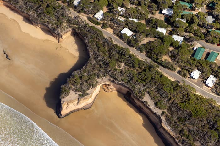 Aerial view of coastal cliffs, sandy beach, surf, clifftop vegetation and nearby homes along the Anglesea coastline in Victoria.