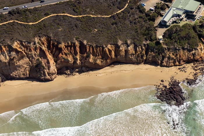 Aerial view of Demons Bluff Beach at Anglesea VIC, showing waves washing onto a narrow sandy beach below eroded sandstone cliffs, with clifftop homes and a coastal road above.