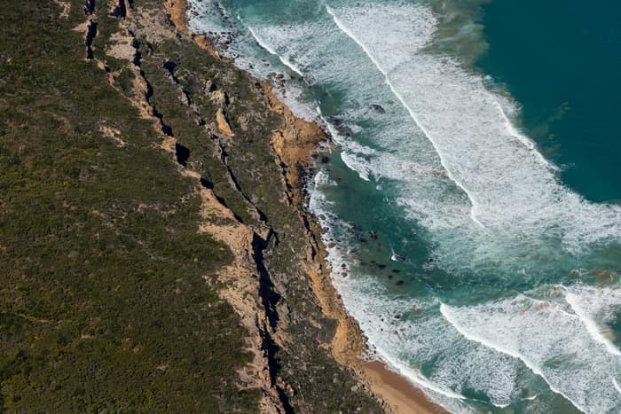 Aerial view of rugged cliffs, coastal scrub, surf and a narrow sandy beach in Port Campbell National Park near Princetown, Victoria.