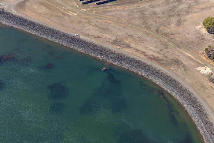 Aerial view of Wurdee Boluc Reservoir shoreline showing green water, a rock-armoured embankment, dry access tracks and vehicles near Wurdiboluc, Victoria.