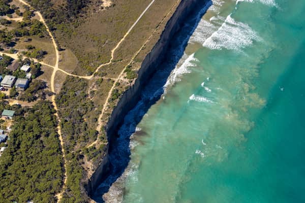 Aerial view of Anglesea cliffs beside Bass Strait, with turquoise water, breaking waves, coastal scrub, walking tracks and nearby houses in Anglesea VIC.