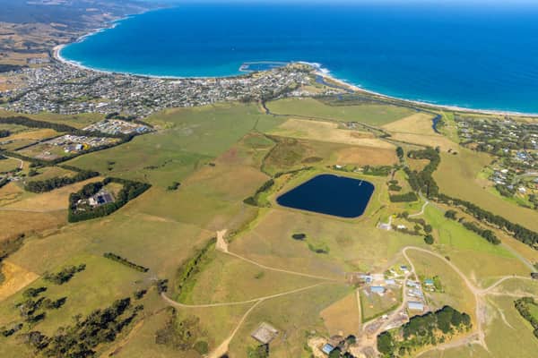 Aerial view of Apollo Bay township and harbour beside the curved coastline, with farmland, a water storage pond and residential areas in Colac Otway, Victoria.