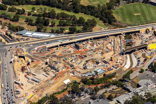 Aerial view of the North East Link construction site beside Manningham Road in Bulleen, with cranes, bridge works, traffic, parkland and a nearby sports oval visible.