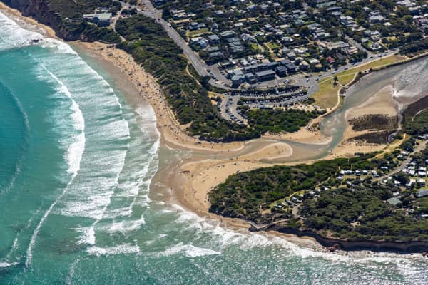 Aerial view of the Anglesea River mouth meeting the surf at Anglesea Beach, with Great Ocean Road, township streets, car parks and coastal vegetation in Anglesea, Victoria.