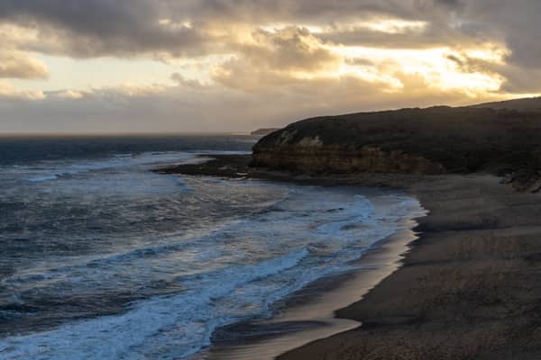 Aerial view of Bells Beach coastline in Bells Beach VIC, showing surf rolling onto the beach below coastal cliffs under a cloudy golden hour sky.